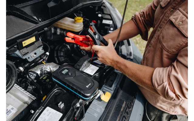 Persoon met opgerolde mouwen houdt een rode startkabelklem boven een autobatterij onder een open motorkap