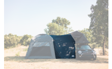 Small black car with an attached gray tent extension on a sunlit field, trees in the background