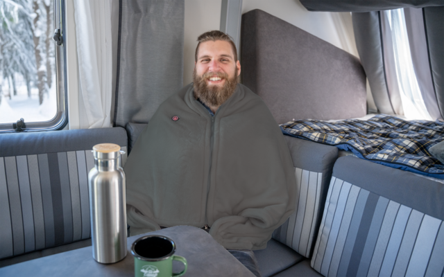 Bärtiger Mann in grauer Decke sitzt im Inneren eines Wohnmobils; Thermosflasche und Tasse auf dem Tisch; Schnee draußen am Fenster sichtbar