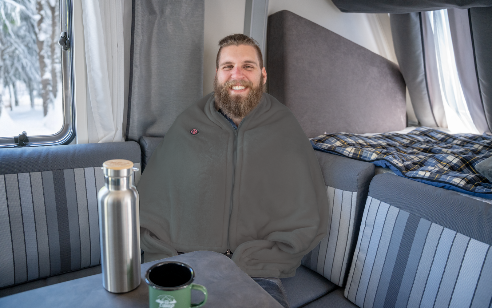 Bärtiger Mann in grauer Decke sitzt im Inneren eines Wohnmobils; Thermosflasche und Tasse auf dem Tisch; Schnee draußen am Fenster sichtbar