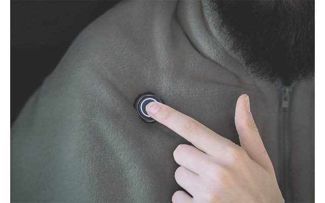 Nahaufnahme: Eine Hand mit Zeigefinger drückt einen runden, beleuchteten Knopf auf der Schulter einer Person; Teil von Hals und Ohr sowie eine dunkle Jacke sichtbar