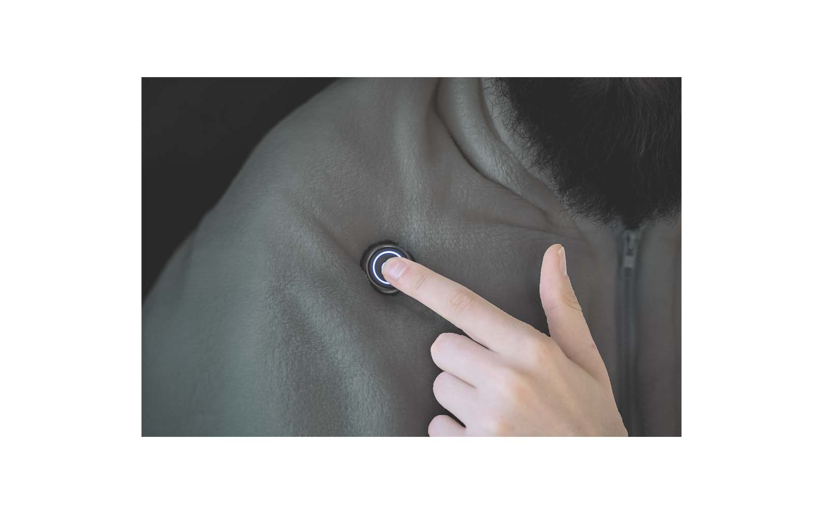 Nahaufnahme: Eine Hand mit Zeigefinger drückt einen runden, beleuchteten Knopf auf der Schulter einer Person; Teil von Hals und Ohr sowie eine dunkle Jacke sichtbar