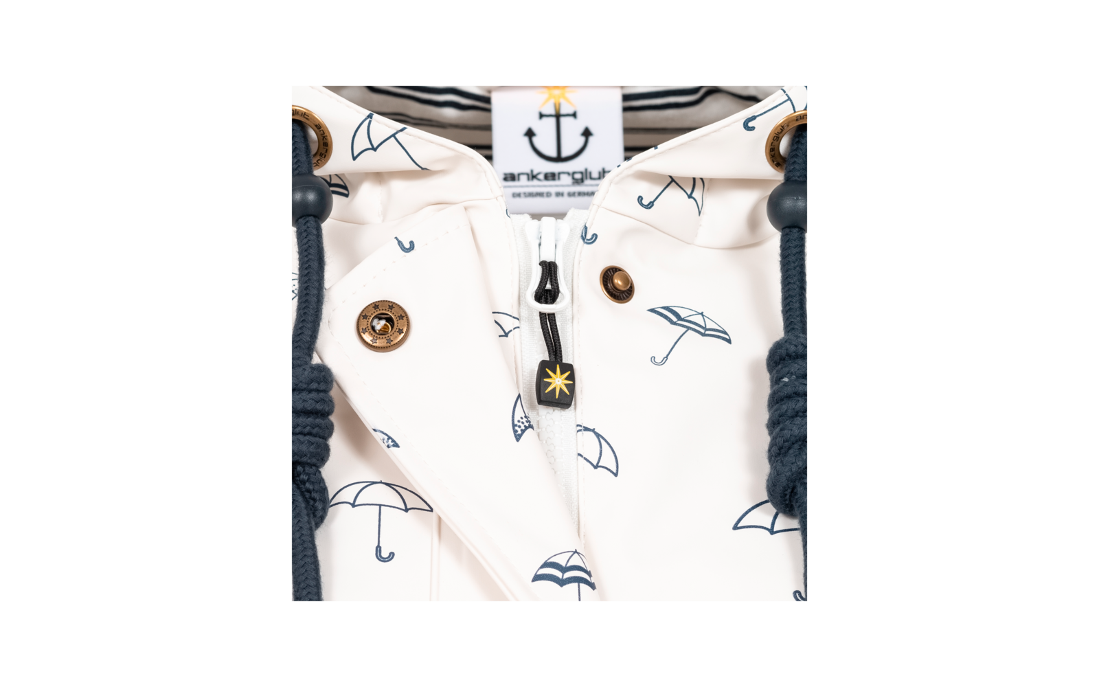 Close-up van een wit kledingstuk met blauwe paraplu‑motieven, zichtbare rits, label met ankersymbool en tekst, goudkleurige drukknopen en donkerblauwe koorden met metalen ringen