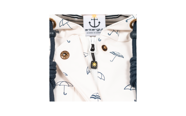 Close-up van een wit kledingstuk met blauwe paraplu‑motieven, zichtbare rits, label met ankersymbool en tekst, goudkleurige drukknopen en donkerblauwe koorden met metalen ringen