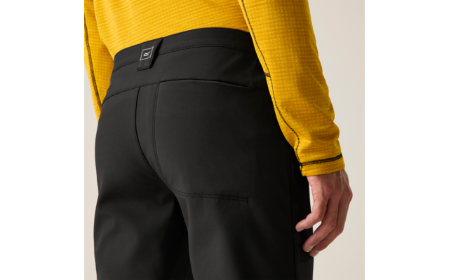 Achteraanzicht van taille tot dij: zwarte broek met stiknaad en klein label in de tailleband, geel langarmshirt, rechterhand langs het lichaam