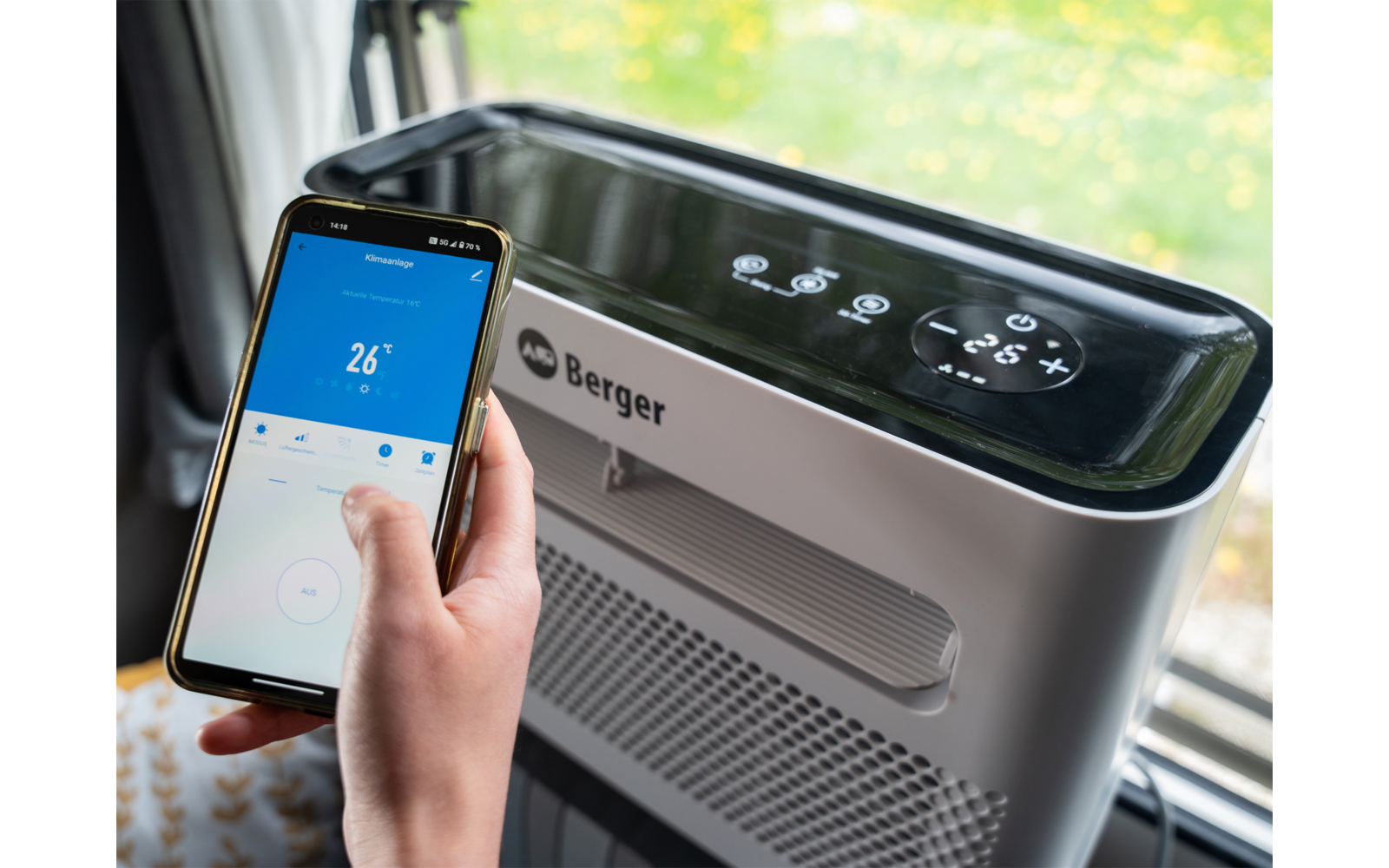 Hand hält Smartphone mit App-Anzeige '26°' vor weißem Gerät mit Berger-Logo und Touchdisplay, Fenster mit grünem Außenbereich im Hintergrund
