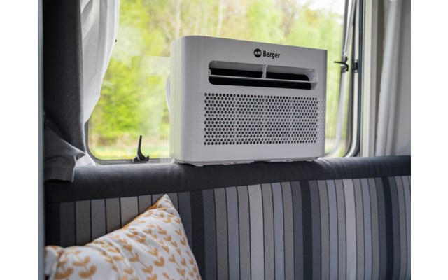 Weißes Gerät mit Lüftungsöffnung und Aufschrift "Berger" auf gepolsterter Rückenlehne vor einem geöffneten Fenster mit Bäumen; gestreiftes Polster und Kissen mit Blattmuster