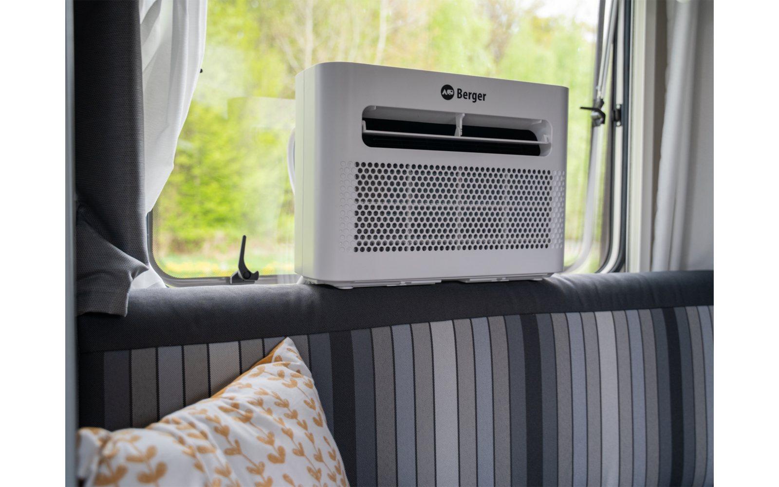Weißes Gerät mit Lüftungsöffnung und Aufschrift "Berger" auf gepolsterter Rückenlehne vor einem geöffneten Fenster mit Bäumen; gestreiftes Polster und Kissen mit Blattmuster