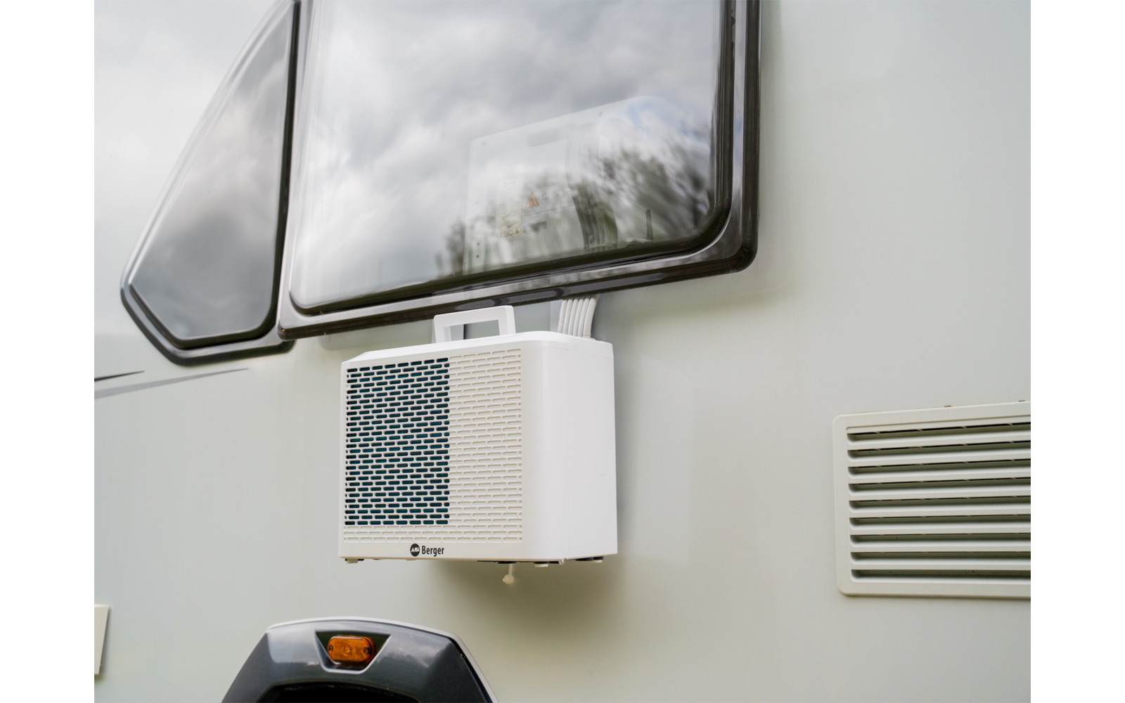 Weißes rechteckiges Gerät mit Gitter an der Außenwand unter einem Fenster; rechts ein Lüftungsgitter; im Fenster Spiegelung von Wolken und Bäumen
