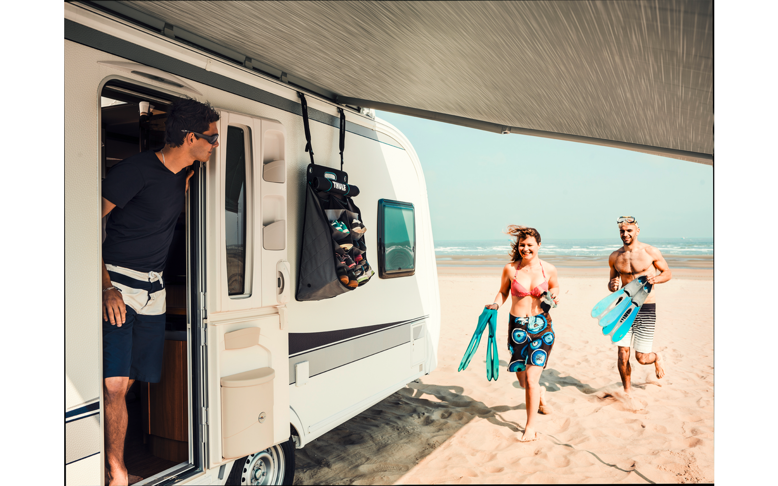 Wohnmobil mit geöffneter Tür auf einem Sandstrand; eine Person steht in der Tür und schaut hinaus; zwei Personen in Bademode laufen am Strand und tragen Schnorchelmasken und Flossen; Meer und Himmel im Hintergrund