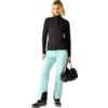 Person mit schulterlangem blondem Haar, schwarze Jacke, helltürkisfarbene Hose, schwarze Stiefel, hält einen schwarzen Helm vor hellgrauem Hintergrund