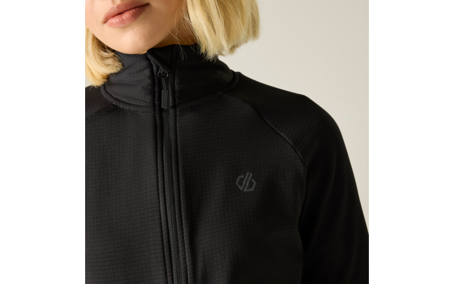 Nahaufnahme des Oberkörpers und des Untergesichts einer Person mit kurzen blonden Haaren, die eine schwarze Jacke mit Reißverschluss und ein kleines graues Logo auf der Brust trägt; heller beiger Hintergrund