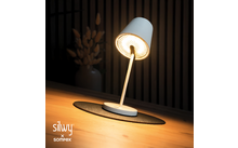 Lampe de bureau blanche avec abat-jour orienté vers le bas, allumée, posée sur un tapis rond noir sur une surface en bois ; en arrière-plan un vase avec des fleurs séchées devant des lattes de bois verticales