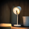 Weiße Schreibtischlampe eingeschaltet neben aufgeschlagenem Buch und weißer Tasse auf Holztisch vor unscharfer Holzverkleidung