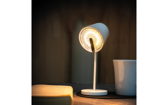 Weiße Schreibtischlampe eingeschaltet neben aufgeschlagenem Buch und weißer Tasse auf Holztisch vor unscharfer Holzverkleidung