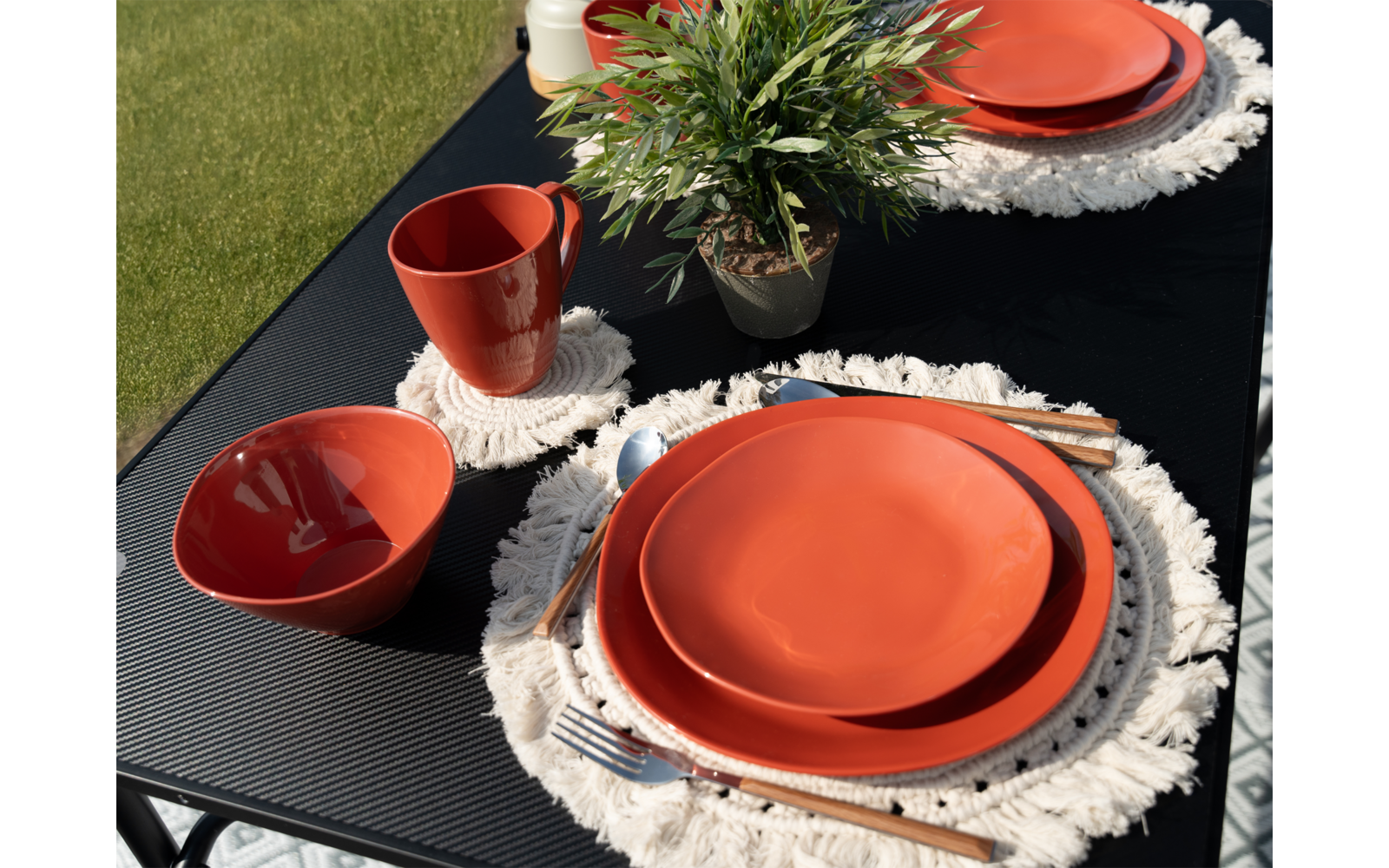 Ein gedeckter Tisch mit orangenen Tellern, Tassen und weißen Häkeluntersetzern auf einer schwarzen Tischdecke, im Hintergrund grünes Gras