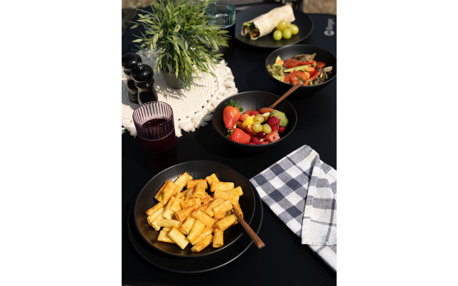 Teller mit Pasta, daneben eine Serviette und ein Glas Rotwein auf einem Holztisch, im Hintergrund Kräuter und Knoblauch