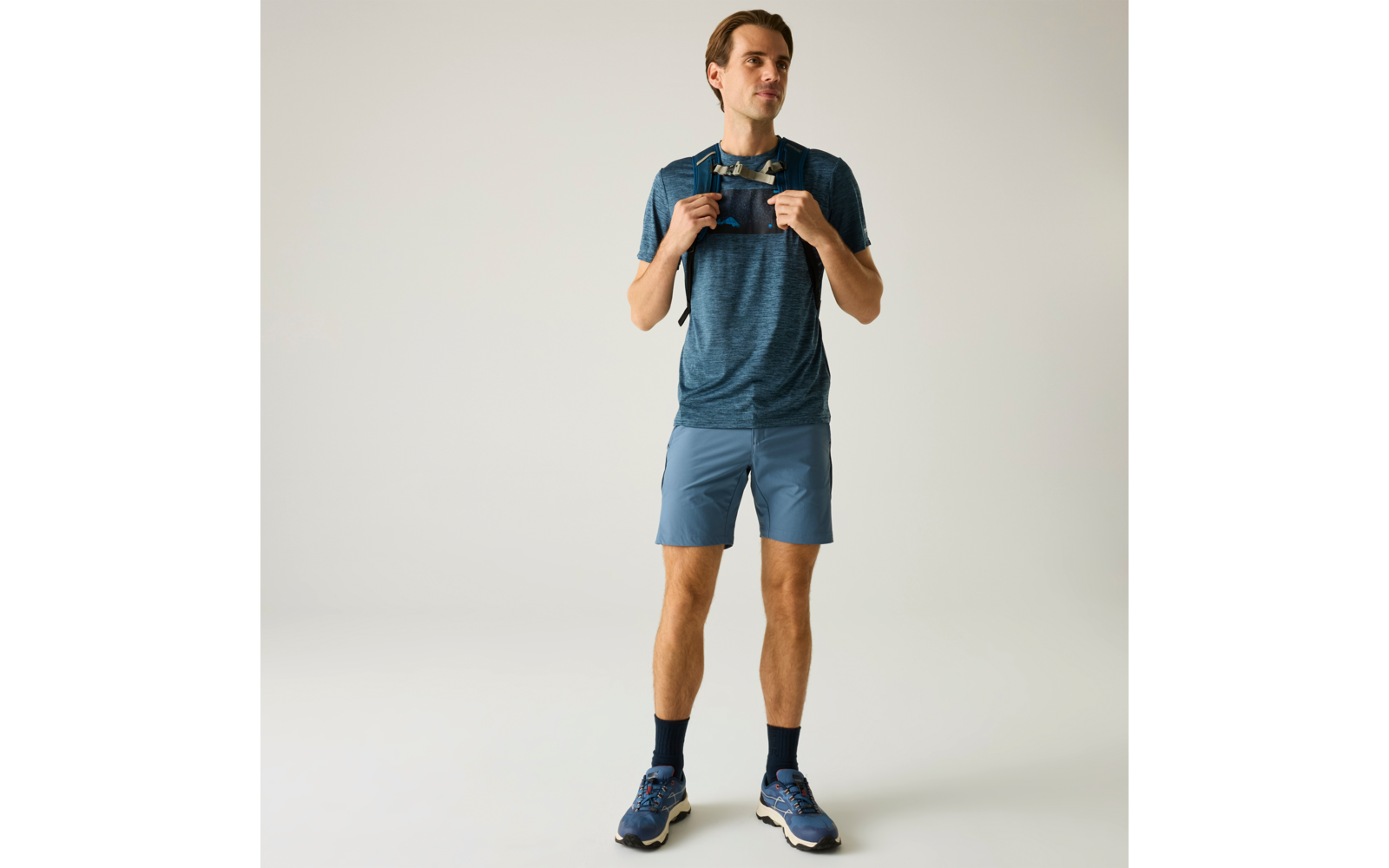 Person steht, trägt blaues Sportshirt, blaue Shorts, dunkle Socken und blaue Laufschuhe, hält eine Sonnenbrille mit beiden Händen vor der Brust, einfarbiger heller Hintergrund