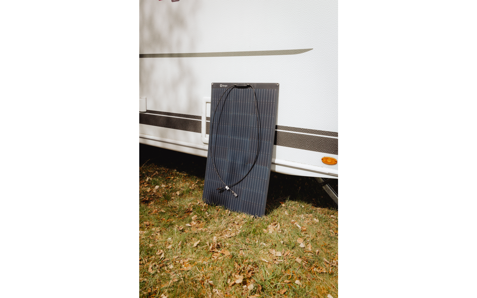 Un pannello solare appoggiato al fianco di una caravan bianca su erba con foglie cadute; cavi neri poggiati sul pannello; la fiancata della caravan mostra strisce orizzontali, un riflettore arancione e ombre di rami