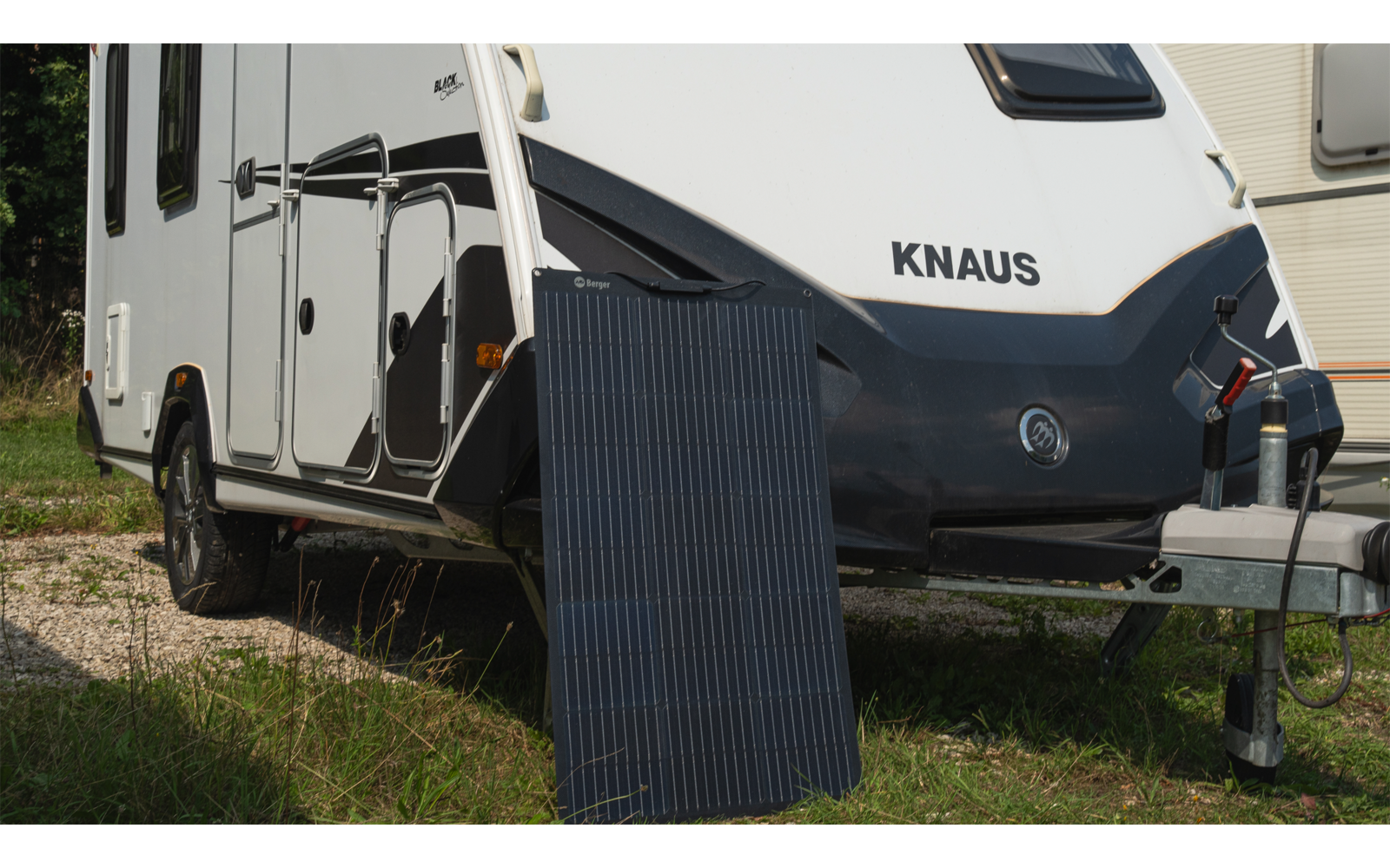 Parte frontal de una caravana blanca con la palabra "KNAUS", un panel solar apoyado delante, hierba y grava en primer plano, enganche de remolque visible