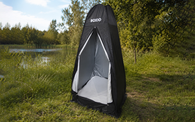 Schwarzes Pop-up-Zelt mit offenem Eingang, silbergrauer Innenauskleidung und Markenaufdruck 'BOXIO', auf Gras neben einem See mit Bäumen und blauem Himmel