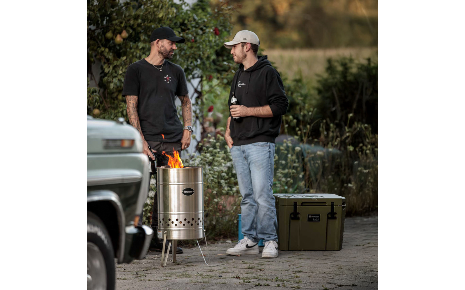 Zwei Personen stehen draußen neben einer brennenden Metallfeuerstelle; eine Person hält eine Flasche; eine grüne Kühlbox und ein geparktes Auto sind in der Nähe; Bäume und Sträucher im Hintergrund