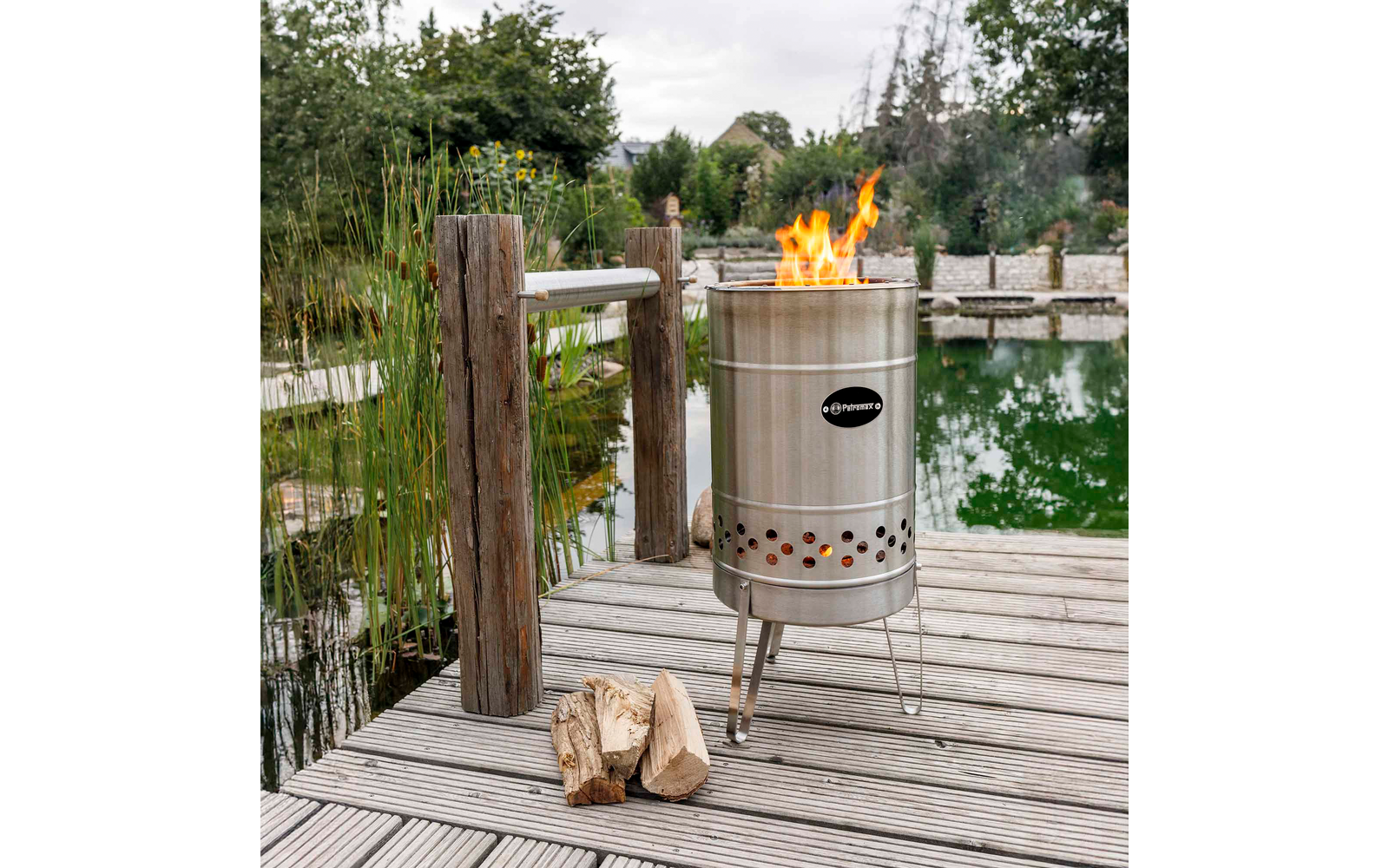 Metallischer, zylindrischer Feuerkorb mit sichtbaren Flammen auf einem Holzsteg neben einem Teich; zwei Holzscheite davor; Holzpfosten, Schilf und Bäume im Hintergrund