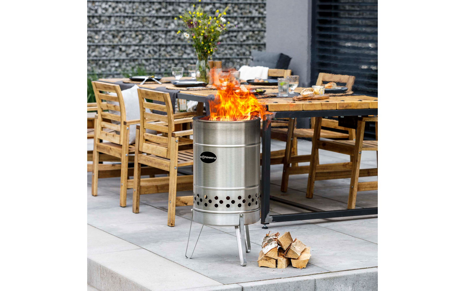 Stählerner Feuerkorb mit Flammen neben einem Holzstapel auf einer Terrasse; im Hintergrund gedeckter Holztisch mit Stühlen und einer Vase mit Blumen