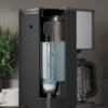 Machine à café noire avec compartiment avant ouvert contenant un récipient cylindrique transparent avec un liquide bleu et un tube spiralé visible ; verseuse en verre à droite ; tasse blanche et plante en pot verte à gauche