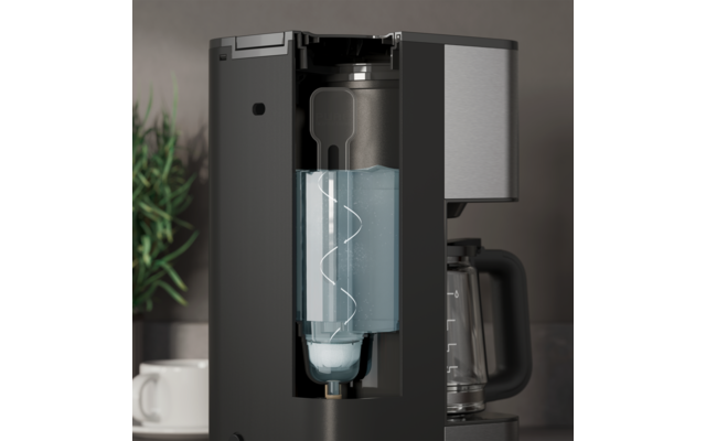 Machine à café noire avec compartiment avant ouvert contenant un récipient cylindrique transparent avec un liquide bleu et un tube spiralé visible ; verseuse en verre à droite ; tasse blanche et plante en pot verte à gauche