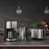 Cafetière avec verseuse en verre, bouilloire électrique et grille‑pain en acier inoxydable sur un plan de travail; deux suspensions et arrière‑plan de cuisine flou avec une plante et un panier de fruits