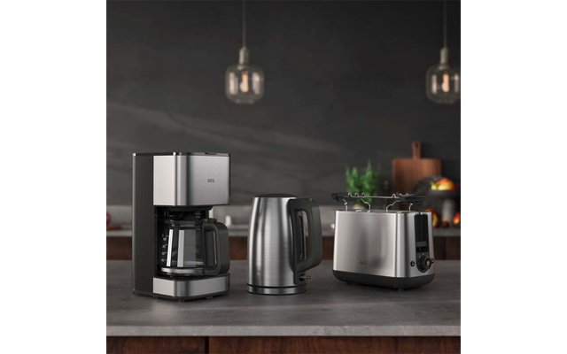 Cafetière avec verseuse en verre, bouilloire électrique et grille‑pain en acier inoxydable sur un plan de travail; deux suspensions et arrière‑plan de cuisine flou avec une plante et un panier de fruits