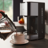 Main versant du café d'une carafe en verre dans une tasse blanche avec soucoupe à côté d'une cafetière gris métallisé sur le plan de travail ; corbeille de fruits floue et fenêtre en arrière-plan
