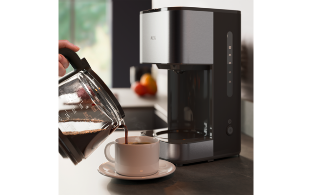 Main versant du café d'une carafe en verre dans une tasse blanche avec soucoupe à côté d'une cafetière gris métallisé sur le plan de travail ; corbeille de fruits floue et fenêtre en arrière-plan