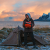 Person wearing a hood stands in front of a pitched tent holding an orange-and-black rolled sleeping bag labeled 'AMBU'; background shows sea, mountains, frost-covered grass and an orange sky