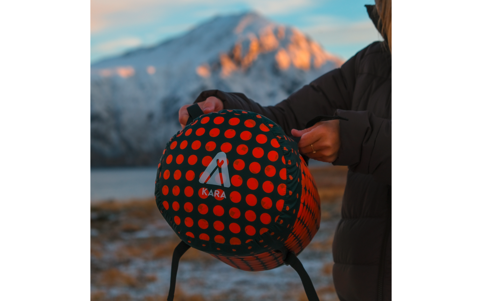Hands of a person holding a round black object patterned with orange dots, a white triangle logo and the text KARA; background shows a lake and snow-covered mountains, the person wears a dark jacket