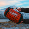 Two hands in a dark jacket hold an orange patterned cylindrical bag with a handle; white text TAMBU connect to nature; snowy mountains and cloudy sky in the background