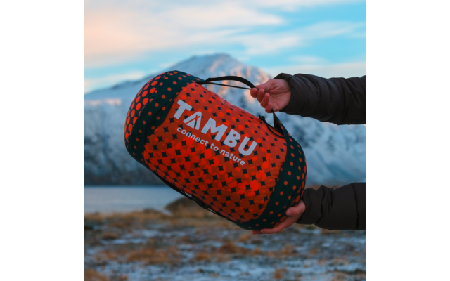 Two hands in a dark jacket hold an orange patterned cylindrical bag with a handle; white text TAMBU connect to nature; snowy mountains and cloudy sky in the background