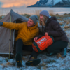 Two people sit in front of a small tent on snowy ground by a shoreline, with water and mountains in the background; one person points with an outstretched arm, the other holds an orange-black sleeping bag labeled TAMBU. They wear winter jackets and beanies