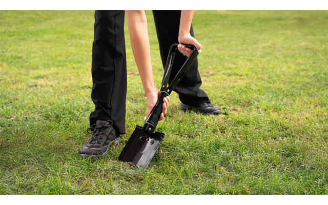 Beine und Hände einer Person in schwarzer Hose und schwarzen Schuhen, die einen Klappspaten mit Metallblatt im grünen Rasen hält