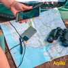 Close-up van een hand die een smartphone boven een geopende kaart houdt. Op de kaart ligt een GPS-apparaat met kabel, daarnaast staan verrekijkers