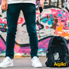 Onderlichaam van een persoon met witte schoenen en blauwe broek voor een kleurrijke graffiti-muur, met een zwarte rugzak op de grond ernaast