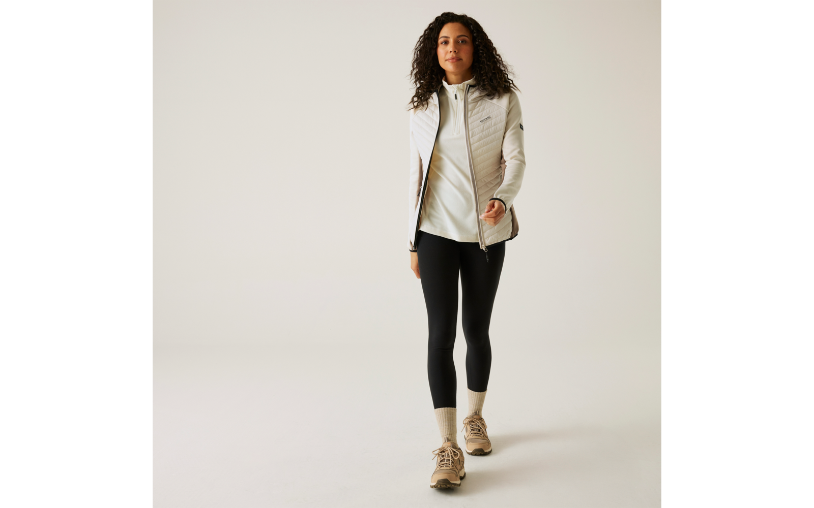 Femme aux cheveux bouclés portant une veste claire, un legging noir et des chaussures claires marchant sur un fond clair