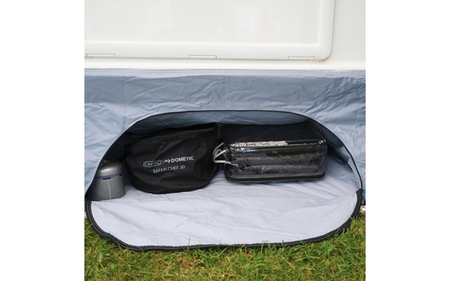 Offenes Fach mit weißer Innenfläche und grauem Stoffrand; darin runde schwarze Tasche mit Aufschrift 'Dometic Safari Chef 30', durchsichtige Reißverschlusstasche mit dunklem rechteckigem Gegenstand und kleiner Metallzylinder; Gras vor dem Fach