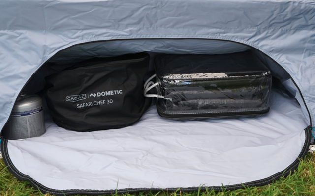 Offenes Fach mit weißer Innenfläche und grauem Stoffrand; darin runde schwarze Tasche mit Aufschrift 'Dometic Safari Chef 30', durchsichtige Reißverschlusstasche mit dunklem rechteckigem Gegenstand und kleiner Metallzylinder; Gras vor dem Fach