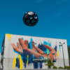 Graffiti an einer Wand mit zwei Händen und bunten Farbstreifen, darüber schwebt ein Fußball vor blauem Himmel