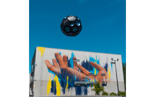 Graffiti an einer Wand mit zwei Händen und bunten Farbstreifen, darüber schwebt ein Fußball vor blauem Himmel