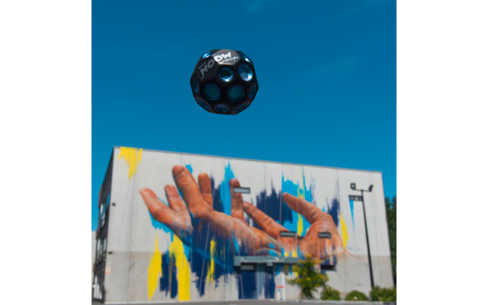 Graffiti an einer Wand mit zwei Händen und bunten Farbstreifen, darüber schwebt ein Fußball vor blauem Himmel