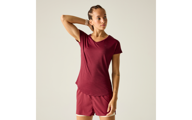 Person vor hellem neutralem Hintergrund, trägt bordeauxrotes kurzärmeliges Shirt und passende Shorts, linke Hand hinter dem Kopf, rechte Hand seitlich, geflochtene Haare, Uhr am linken Handgelenk, kleines Tattoo am rechten Unterarm