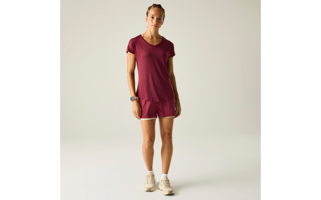 Person steht frontal, trägt bordeauxrotes T‑Shirt und kurze Sportshorts, weiße Sneakers, Smartwatch am Handgelenk, heller einfarbiger Hintergrund